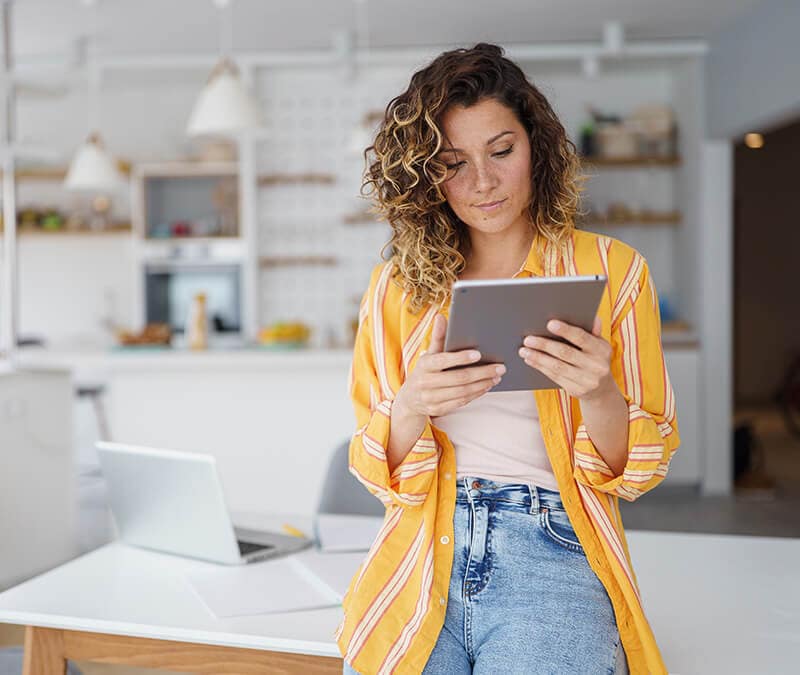 Woman wearing a yellow shirt and leaning against a table, reading her iPad.