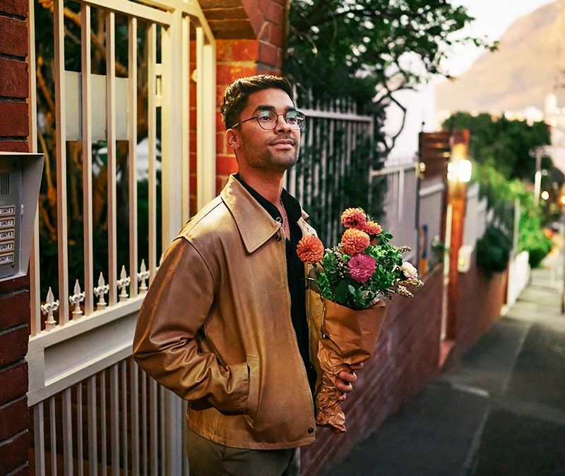 A man waiting outside a brick house, holding a bouquet of flowers.
