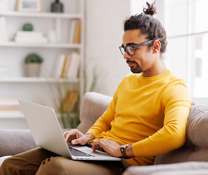 A man in glasses and a yellow shirt sitting on a couch with a laptop, engrossed in his work and wondering whether Macs get viruses.