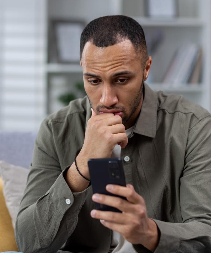 A worried man in a grey shirt is looking at his phone, unsure if it is tapped or hacked.